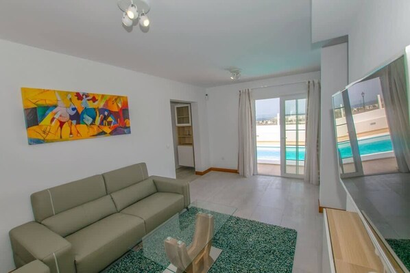 A living room with a sofa, a glass coffee table, a painting, and a view of a pool and beach through large windows.