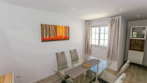 Een moderne eethoek met een glazen tafel, vier stoelen, een schilderij, een raam met gordijnen en een inbouwplank.