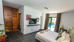 A hotel room with a bed, a TV mounted on the wall, a potted plant, and a view of the outside. - Villa Lanson - Poolside Retreat by Villas Now (Playa Blanca)