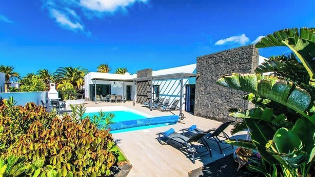 A modern pool area with lounge chairs, a stone wall, and tropical plants. - Villa Lanson - Poolside Retreat by Villas Now (Playa Blanca)