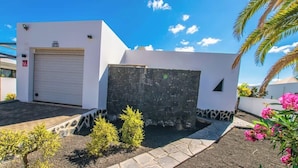 A modern building with a garage door, a stone wall, and a small garden with shrubs and flowers. - Villa Lanson - Poolside Retreat by Villas Now (Playa Blanca)