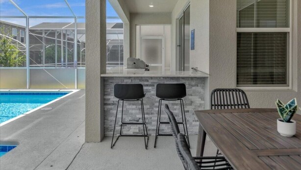 Zona junto a la piscina con un bar, dos taburetes, una mesa y una planta en maceta.