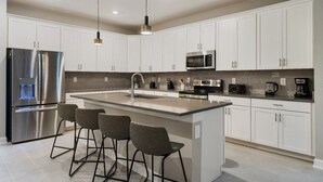 Una cocina moderna con armarios blancos, electrodomésticos de acero inoxidable, una isla central con taburetes altos e iluminación colgante.