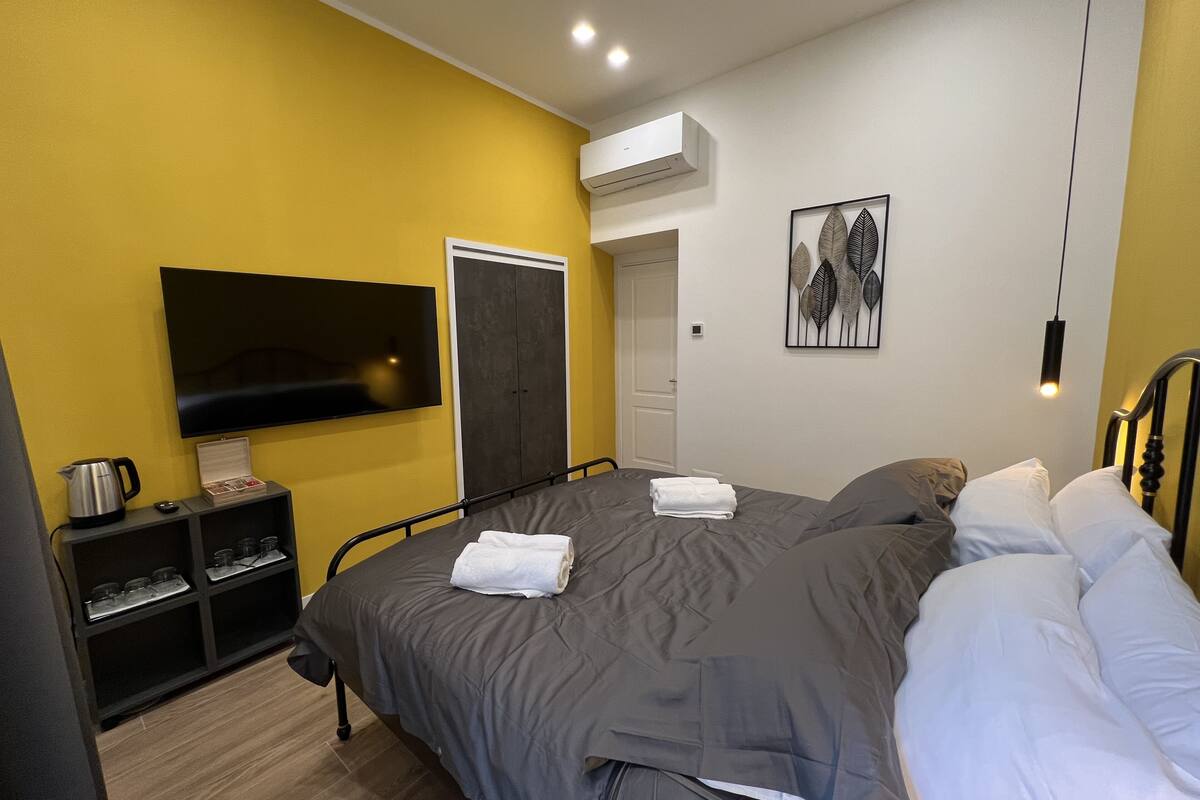 A modern hotel room with a large bed, a flat-screen TV, a wardrobe, and a small kitchenette.