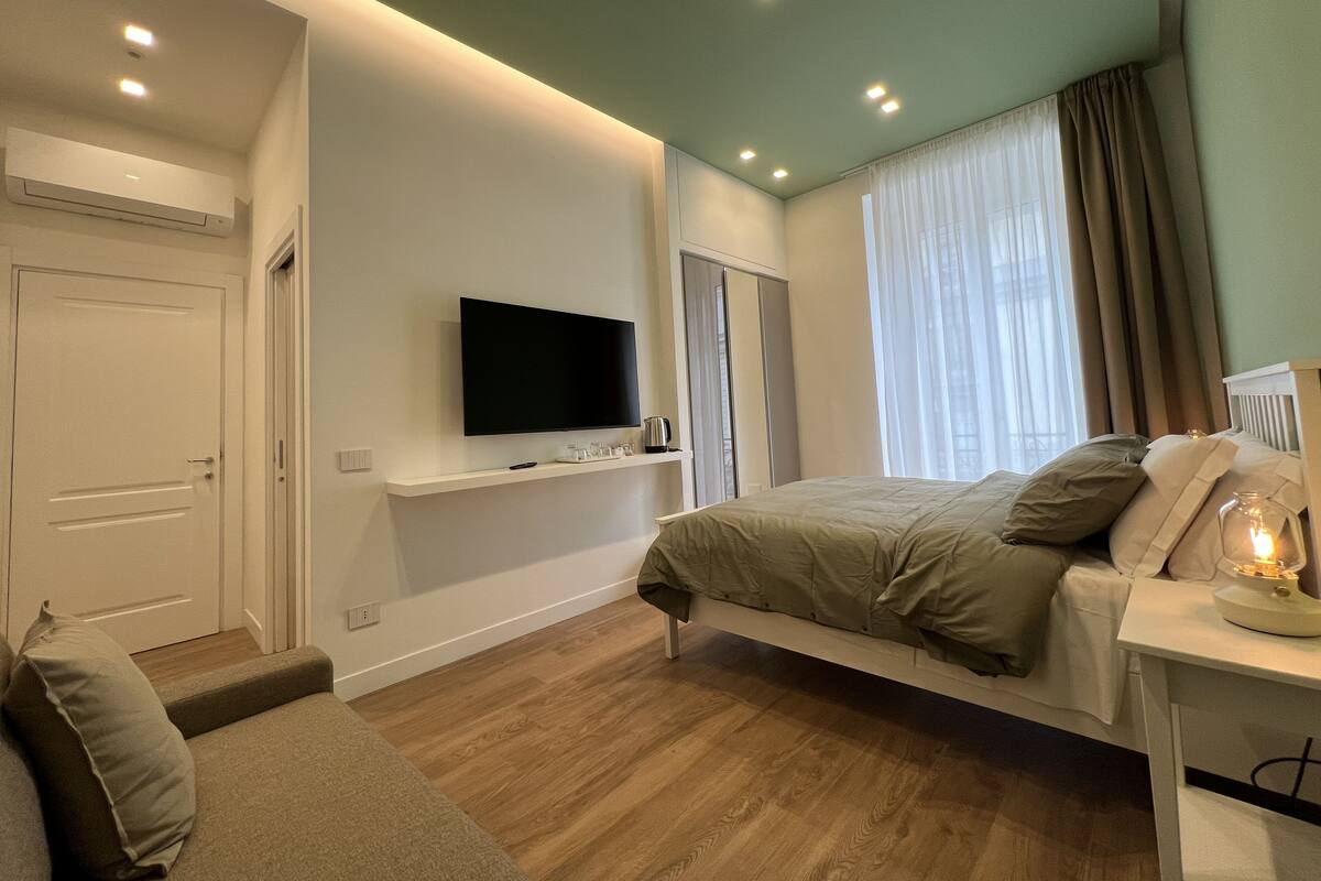 A modern bedroom with a flat-screen TV mounted on the wall, a bed with a headboard, a bedside table with a lamp, and a wooden floor.