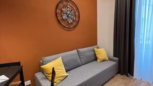Una sala de estar moderna con un sofá gris, cojines amarillos, una mesa de comedor de madera y un reloj fijado a la pared.