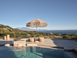 Ein Pool mit Blick auf Meer und Berge, ein gewebter Sonnenschirm und zwei beigefarbene Kissen.