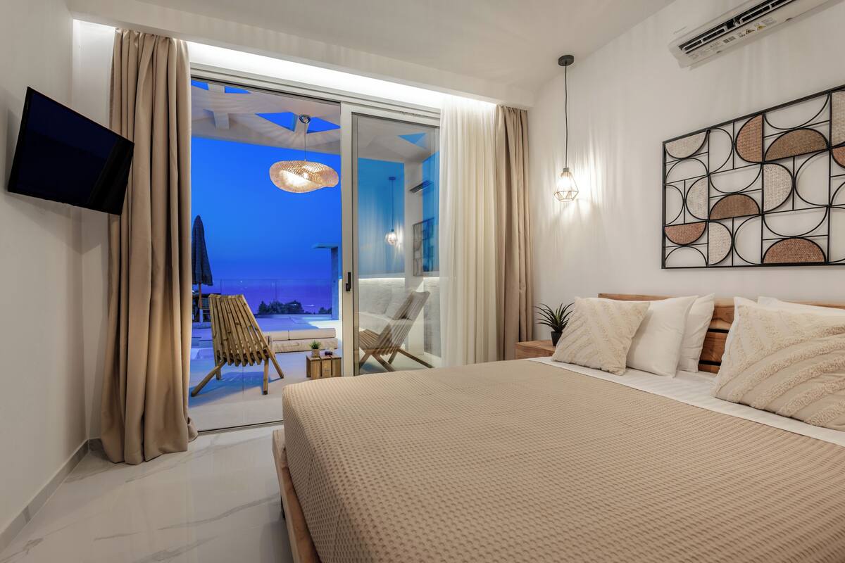 A modern bedroom with a large bed, a flat-screen TV, a ceiling fan, and a view of the beach through the sliding glass door.