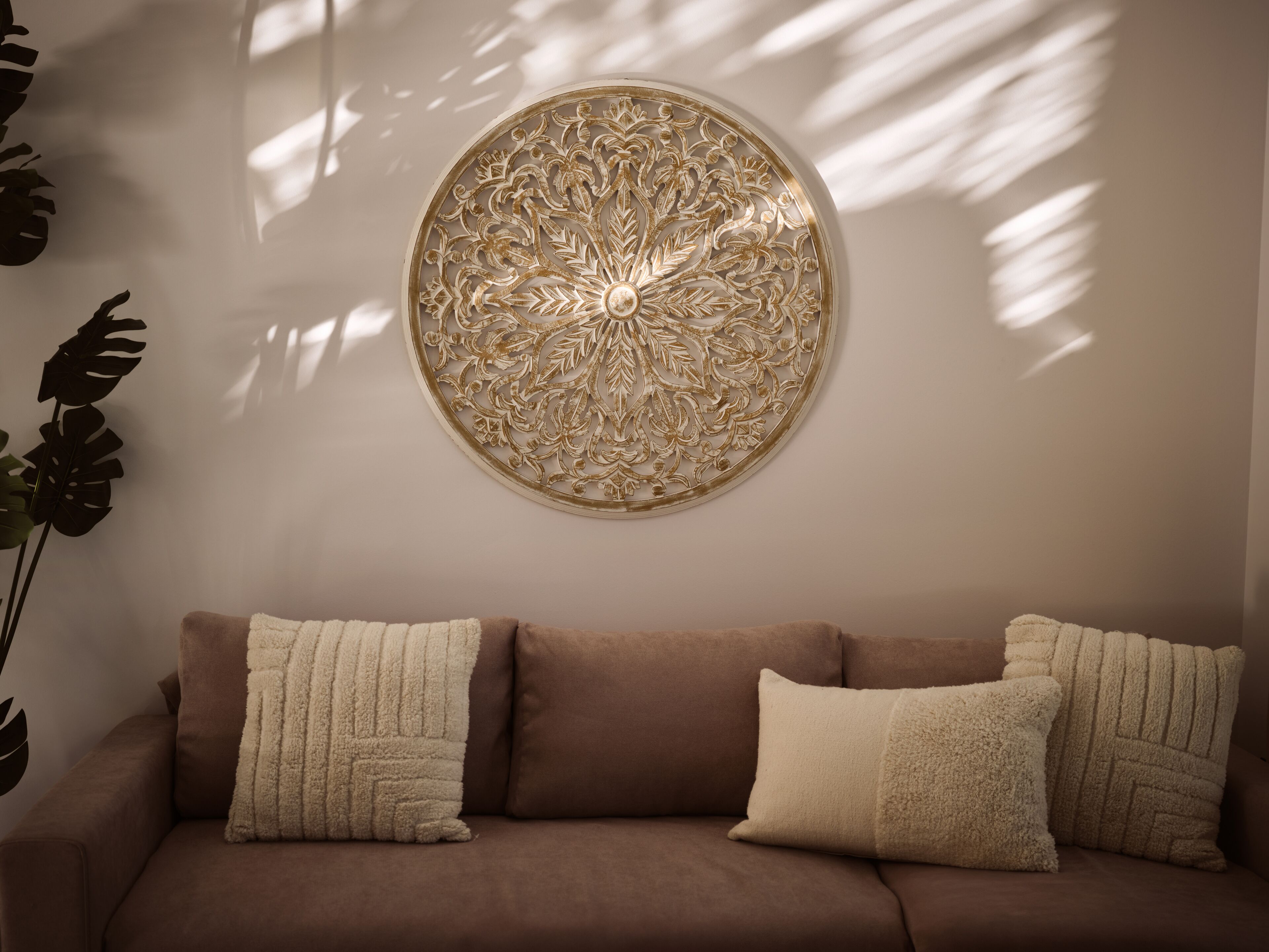Un canapé marron avec trois coussins beige, un élément décoratif rond fixé au mur et une grande plante sur la gauche.