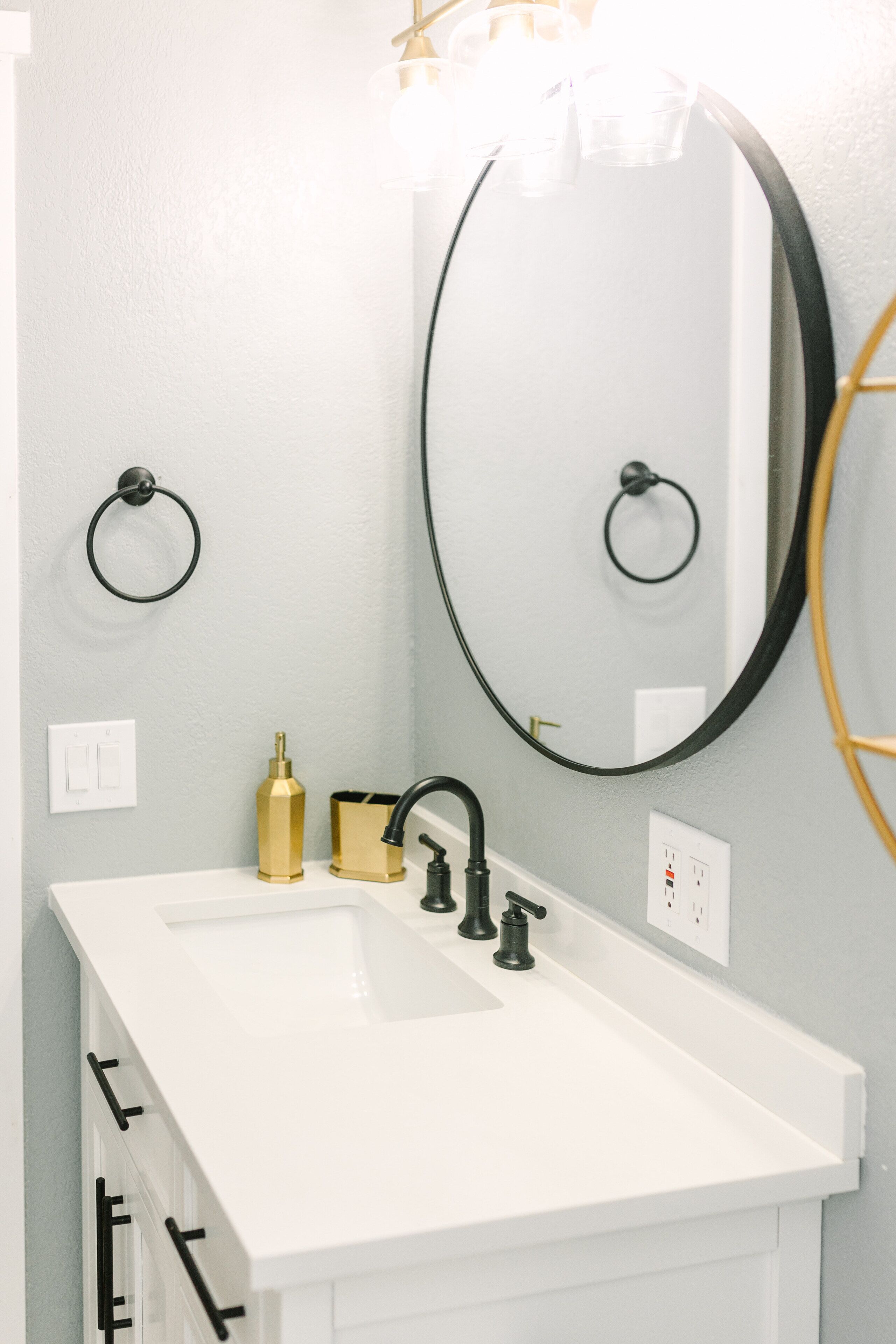 Une salle de bain avec un lavabo blanc, un robinet noir et des accessoires dorés. On y trouve deux porte-serviettes et un miroir avec un cadre noir.