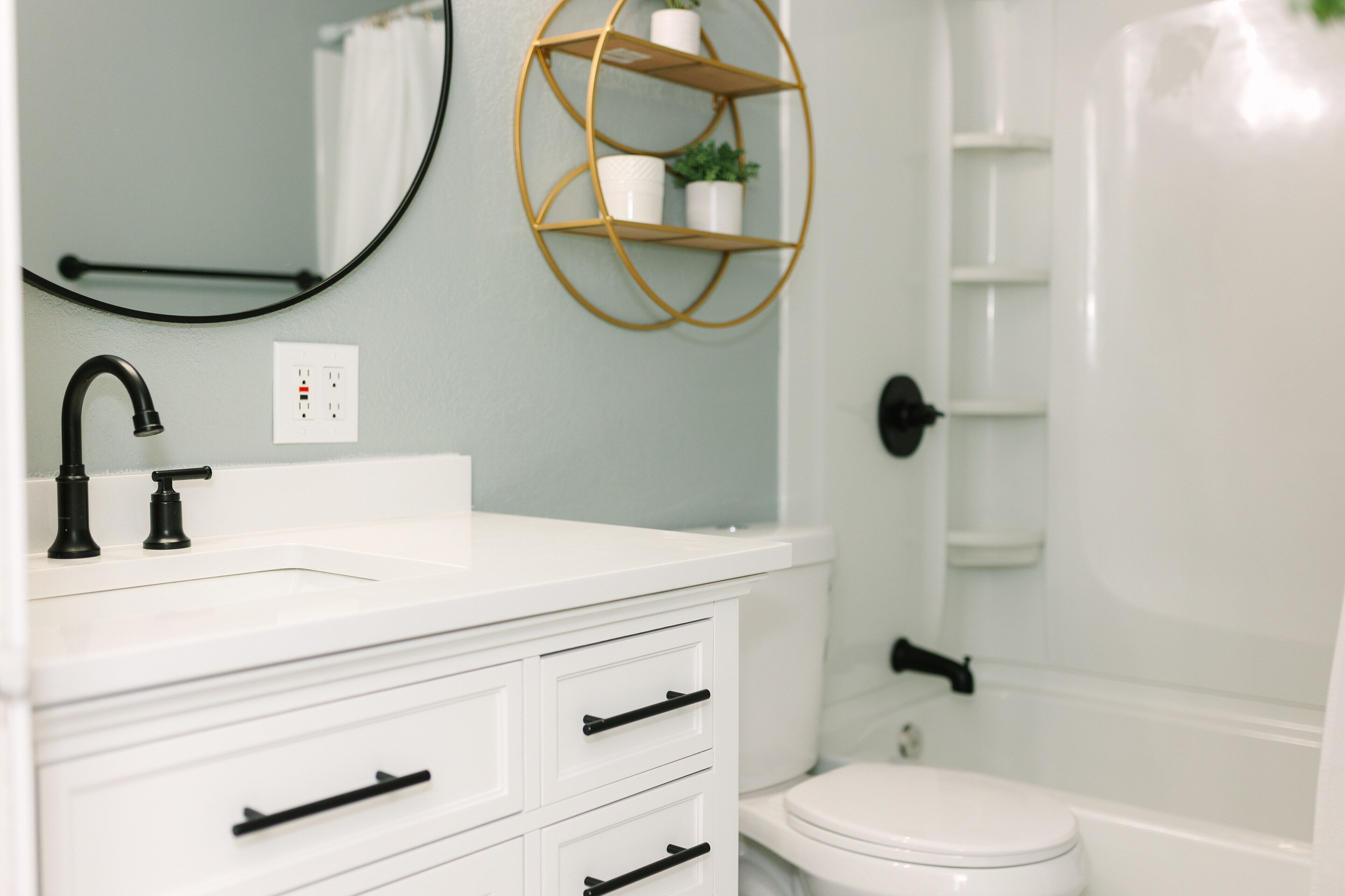 A bathroom with a white sink, black faucet, and a gold-framed shelf with a potted plant.