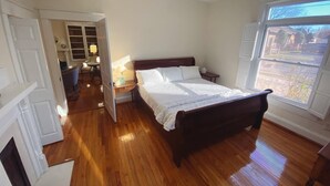 A bedroom with a wooden floor, a bed with white bedding, a nightstand, a lamp, and a window with a view of the outside. - Beautiful historic home in Downtown Murfreesboro-2 bedroom option-sleeps 6 (Murfreesboro)