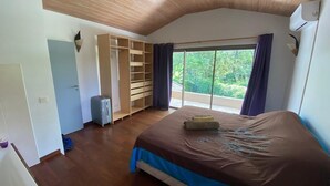 A bedroom with a large bed, wooden ceiling, built-in wardrobe, and a view of greenery through the window.