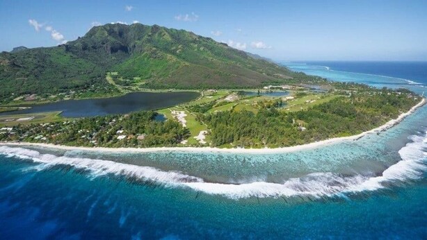 An aerial view of a tropical island with a large mountain, clear blue waters, and a beach.