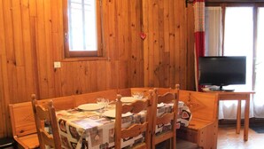 Een kamer met houten lambrisering, een eettafel voor twee personen, een televisie en een raam met gordijnen.