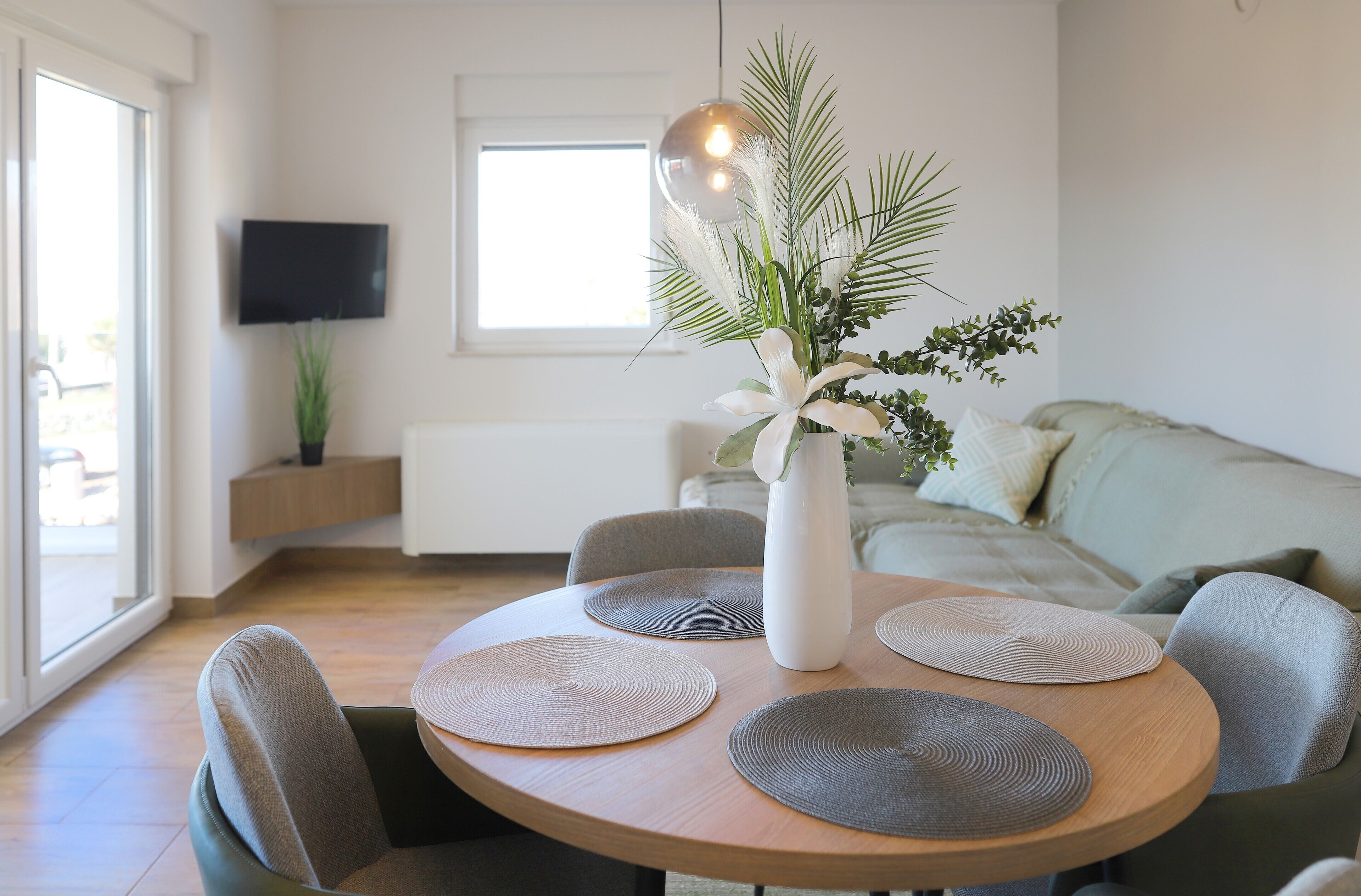Una sala de estar moderna con una mesa de comedor redonda de madera, sillas tapizadas en gris, un jarrón blanco con una planta y un televisor de pantalla plana montado en la pared.