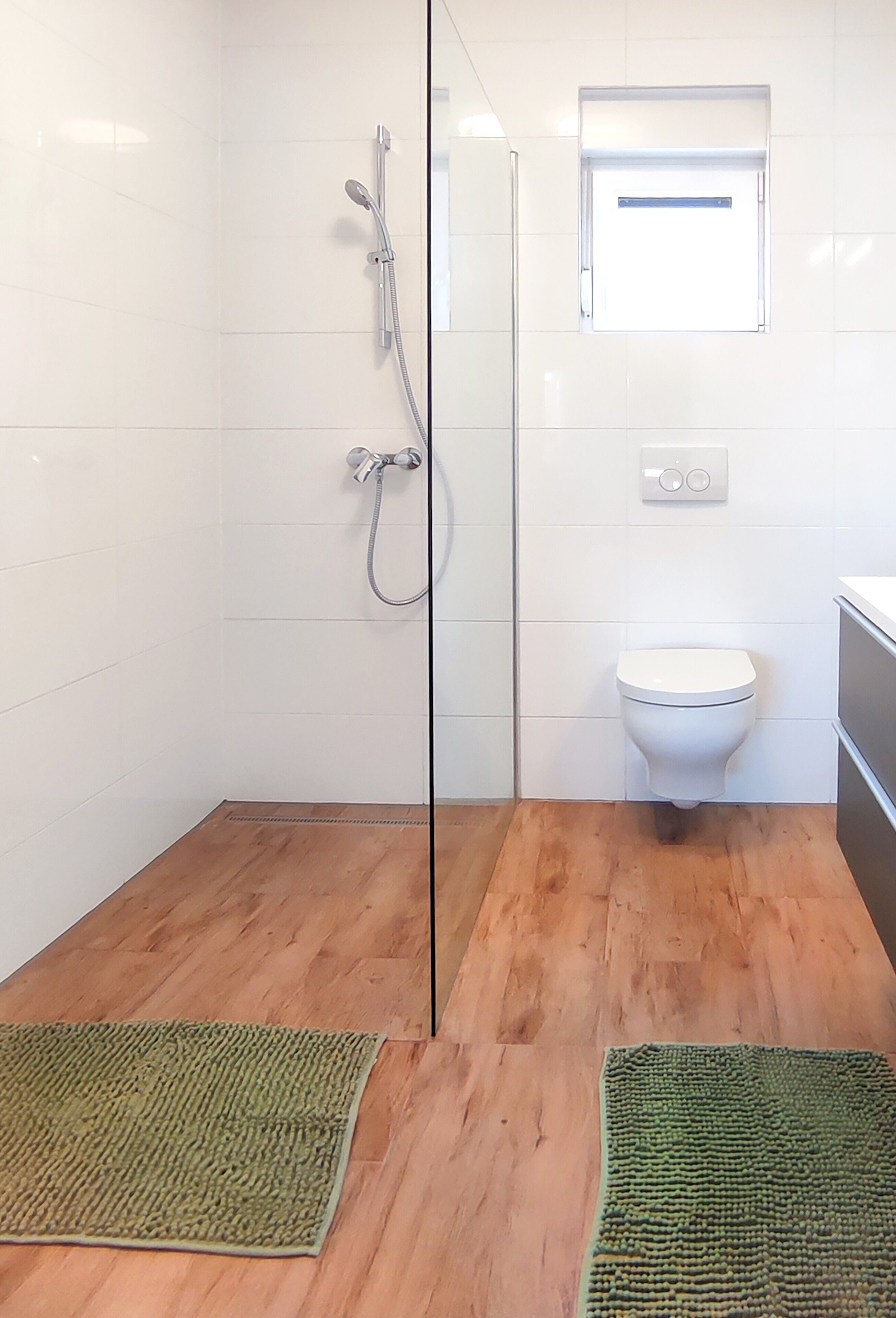 Une salle de bain avec une paroi en verre, une douche, des toilettes et deux tapis verts sur le sol.
