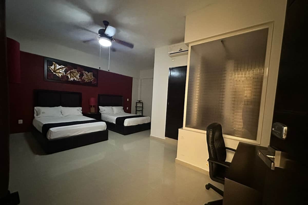 Une chambre d’hôtel avec deux lits, un ventilateur de plafond, une grande fenêtre avec des stores et un bureau avec une chaise.