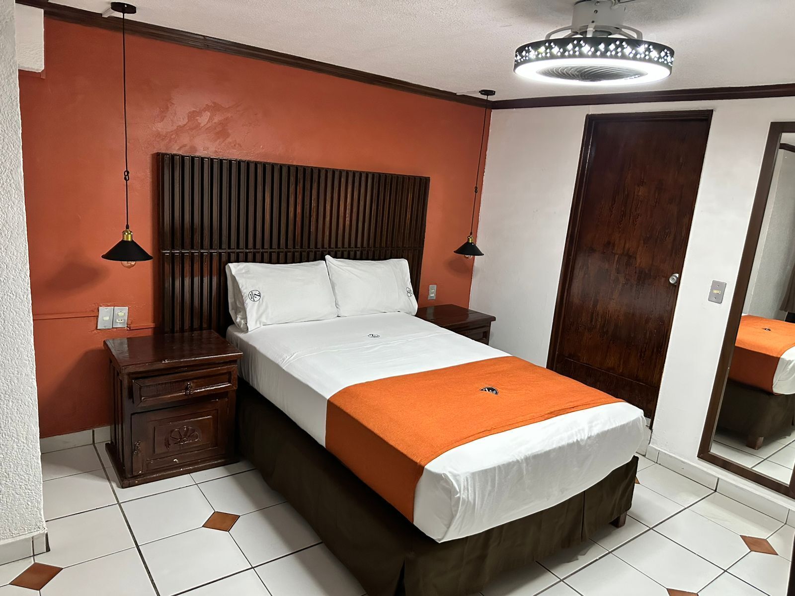 Une chambre d’hôtel avec un lit, des tables de chevet, une tête de lit en bois, un grand miroir et un ventilateur de plafond.