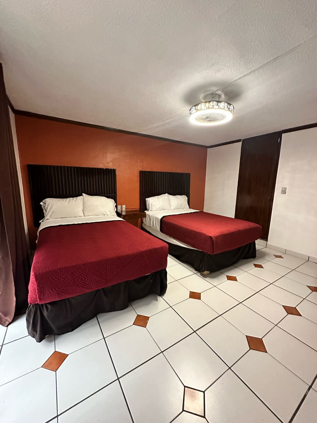 A hotel room with two beds, each with a red bedspread and black skirted legs. The room has a tiled floor with a geometric pattern, a white ceiling with a central light fixture, and a brown accent wall.