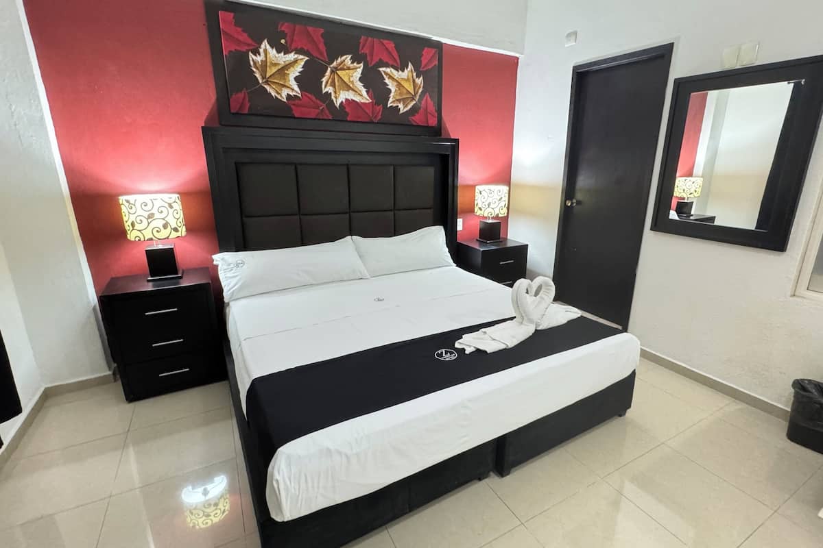 Une chambre d’hôtel bien aménagée, avec un grand lit, deux tables de chevet, un miroir fixé au mur et un motif décoratif en forme de feuilles au-dessus du lit.
