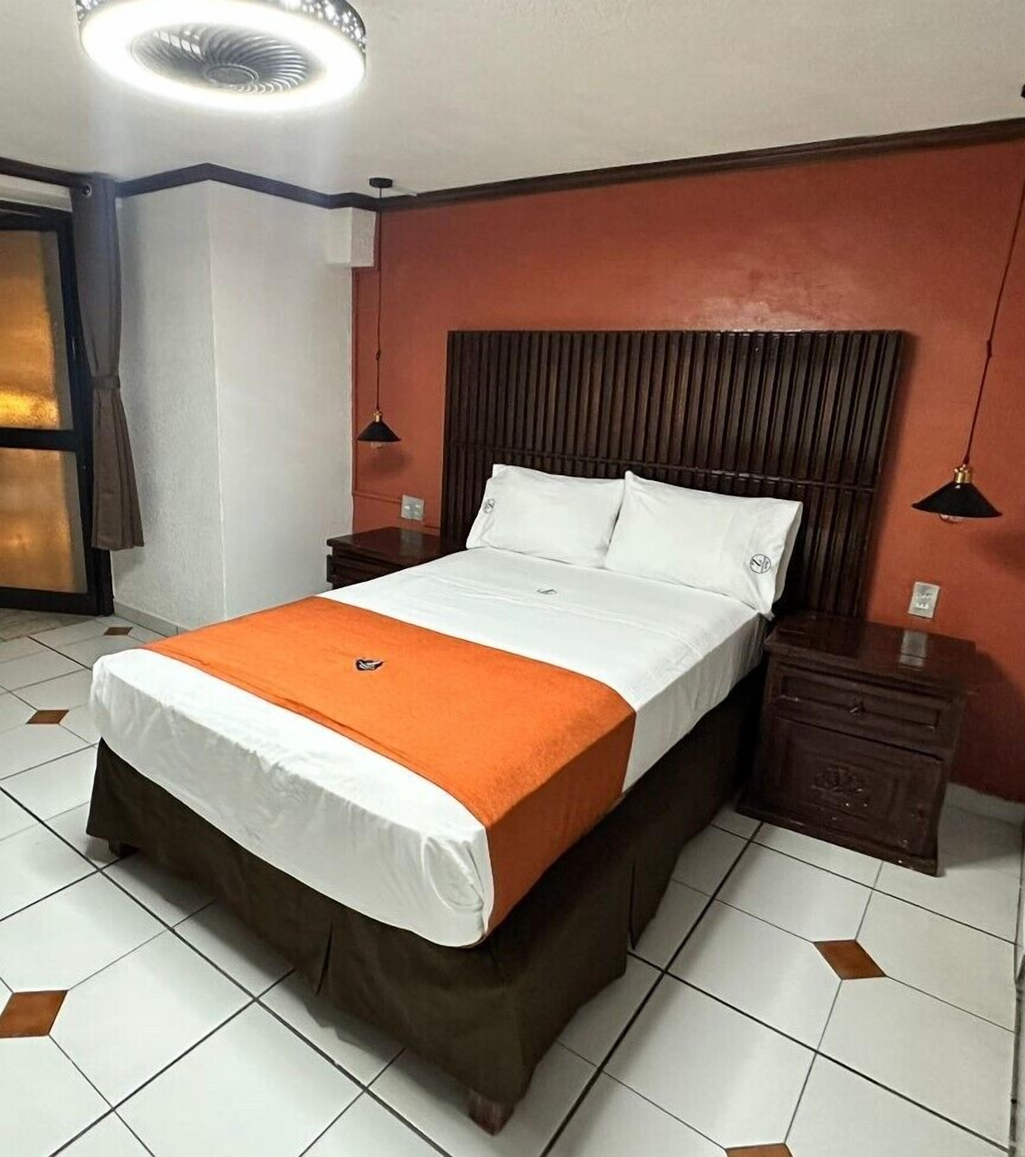 A hotel room with a bed, nightstands, a wooden headboard, a ceiling fan, and a window with curtains.