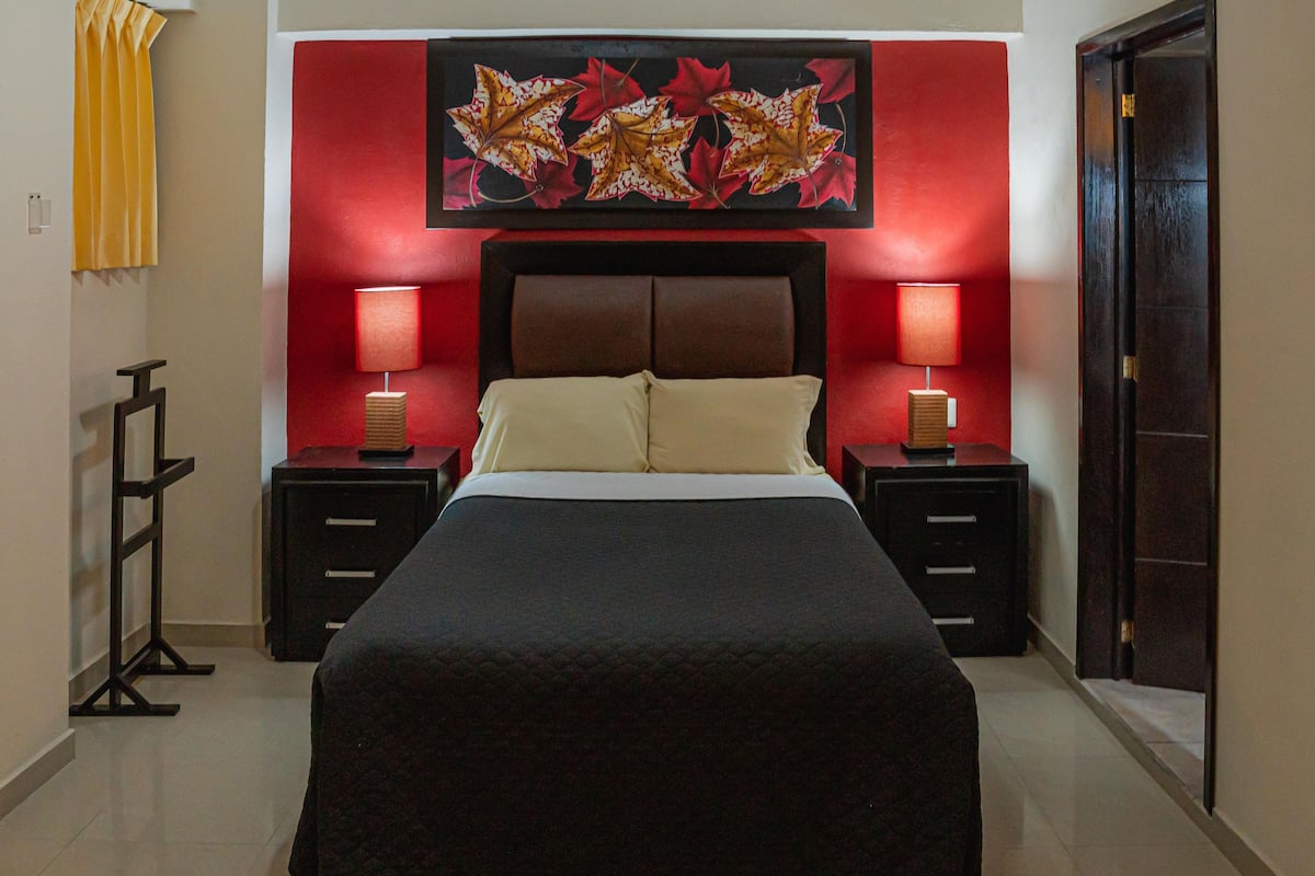 Une chambre d’hôtel avec un grand lit, deux tables de chevet avec des lampes, un mur d’accent rouge orné d’un tableau représentant des feuilles, et une porte sur le côté droit.