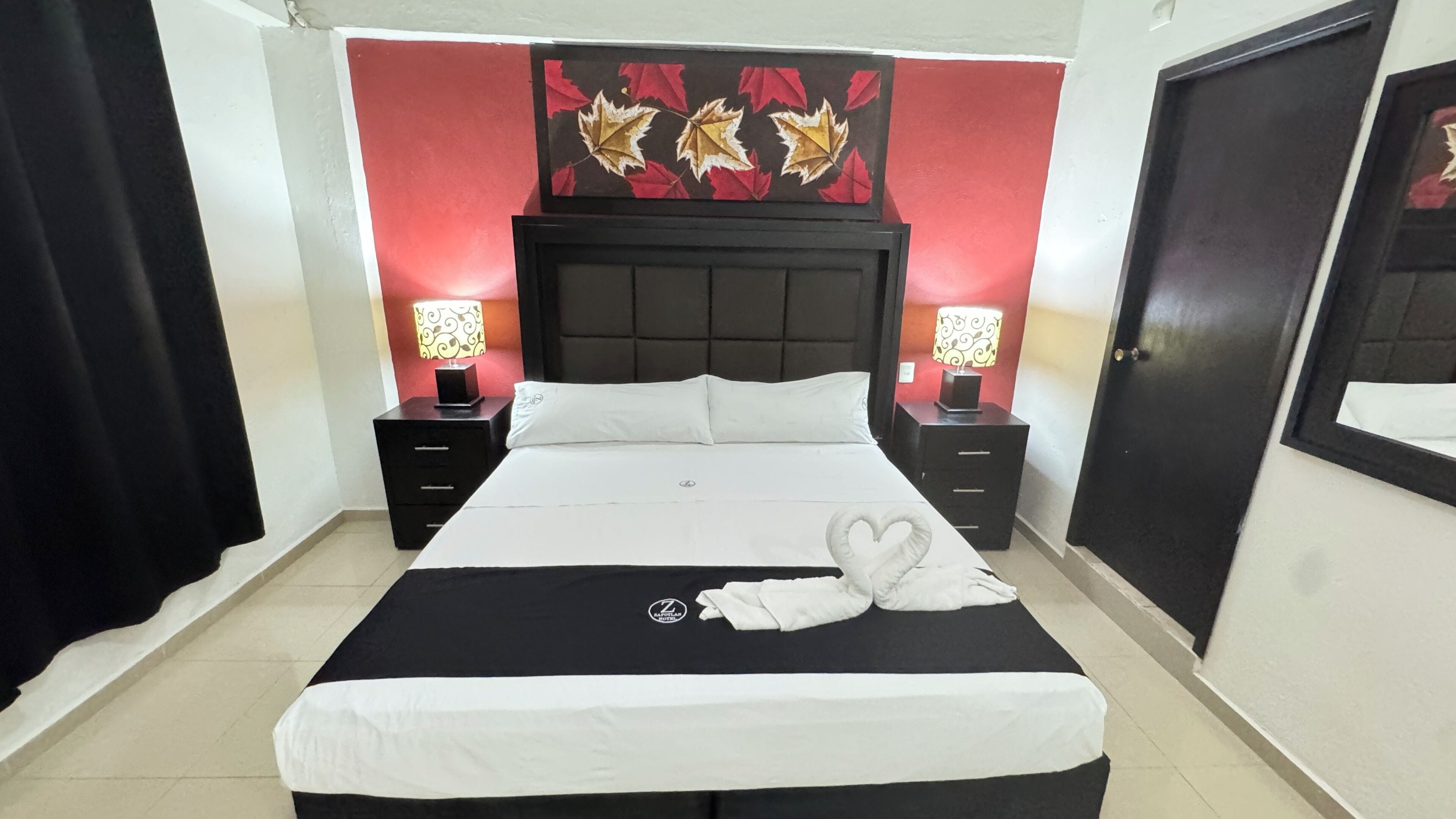 Cama bem arrumada com lençóis brancos, duas mesas de cabeceira com luminárias, parede vermelha e preta com padrão de folhas e uma porta preta.