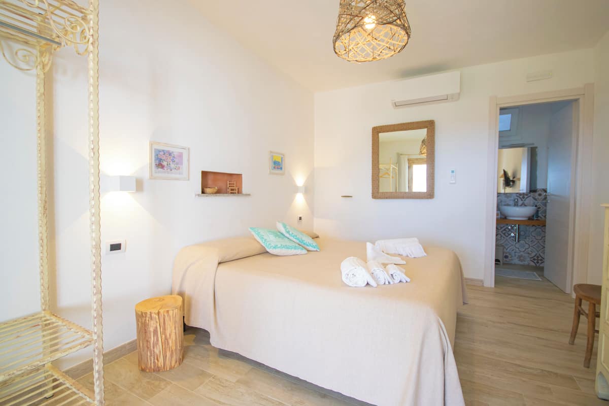 Une chambre à coucher avec un lit, une table de chevet et un miroir. On y trouve également un petit tabouret et une étagère fixée au mur, sur laquelle est posé un bol.