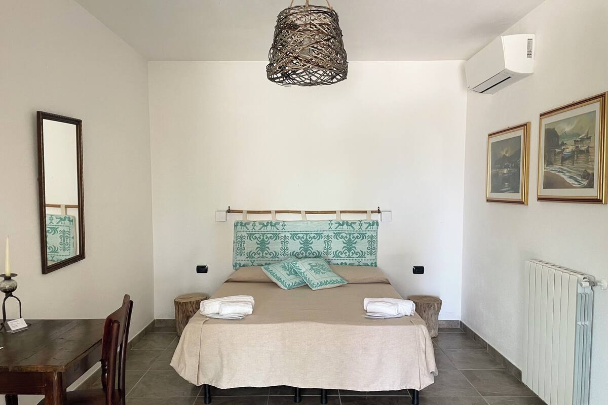 Une chambre à coucher avec un lit, une chaise en bois, une table, un luminaire suspendu, un miroir et deux tableaux encadrés accrochés au mur.
