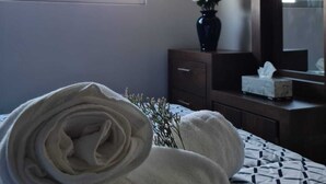 A neatly made bed with a patterned bedspread, a rolled white towel, a vase with dried flowers, and a mirror above a wooden dresser.