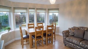 Un comedor con mesa y sillas de madera, ventanas amplias con cortinas y un sofá estampado.