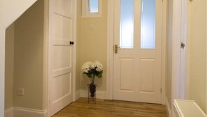 Un pasillo con una puerta blanca, un ventanal pequeño y un jarrón con flores blancas.