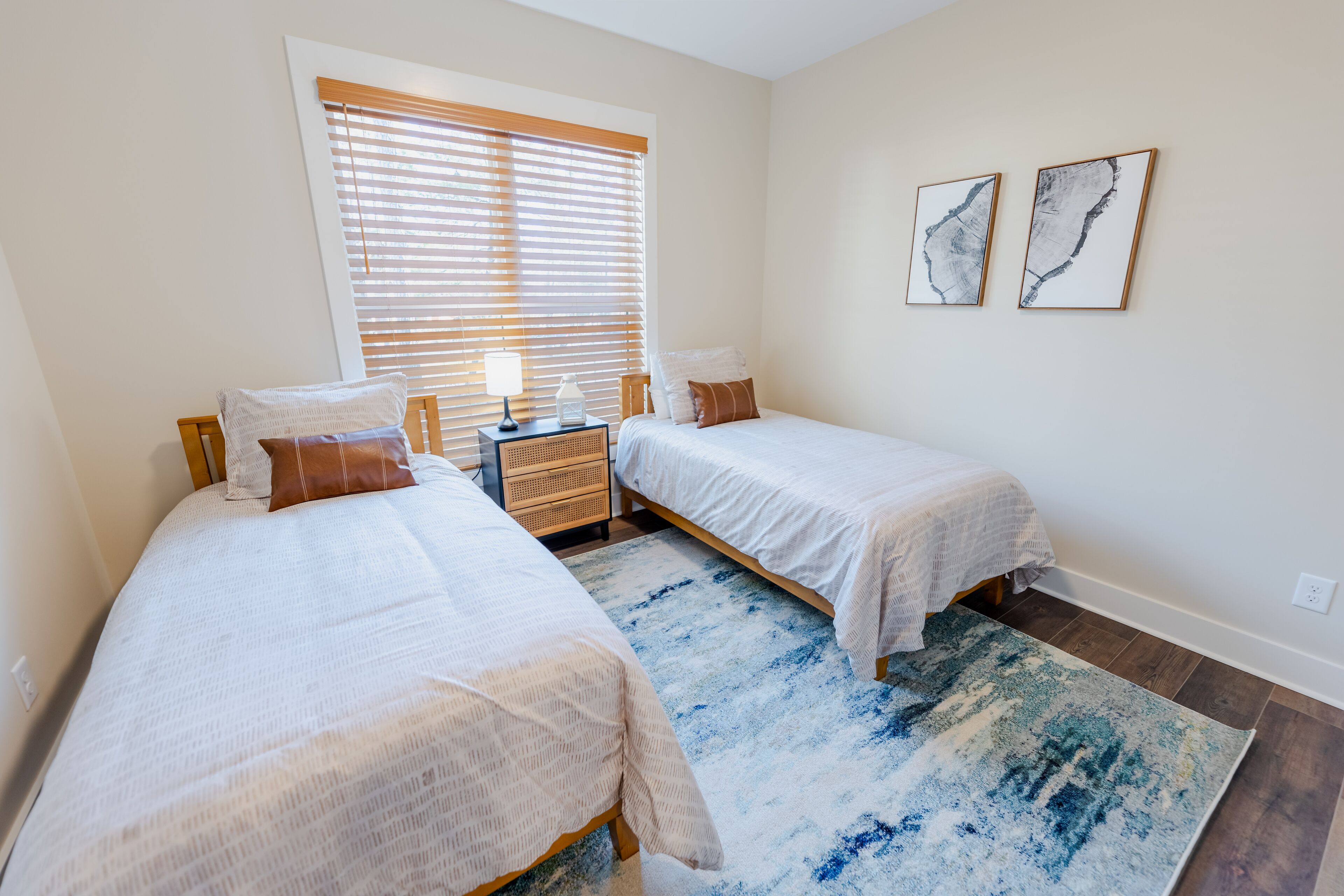 A bedroom with two beds, a nightstand, a window with blinds, and two framed artworks on the wall.