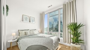 A modern bedroom with a large bed, a small bedside table, a potted plant, and a city view through a large window. - 2 BR Downtown Oasis, AC & stunning views (Vancouver)
