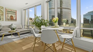 A modern living room with a sofa, coffee table, and dining table with chairs. There are large windows offering a view of a cityscape and a tall building. - 2 BR Downtown Oasis, AC & stunning views (Vancouver)