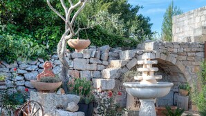 Un jardin avec un mur de pierre, un arbre, un grand bac à fleurs, une fontaine et une vieille charrette.