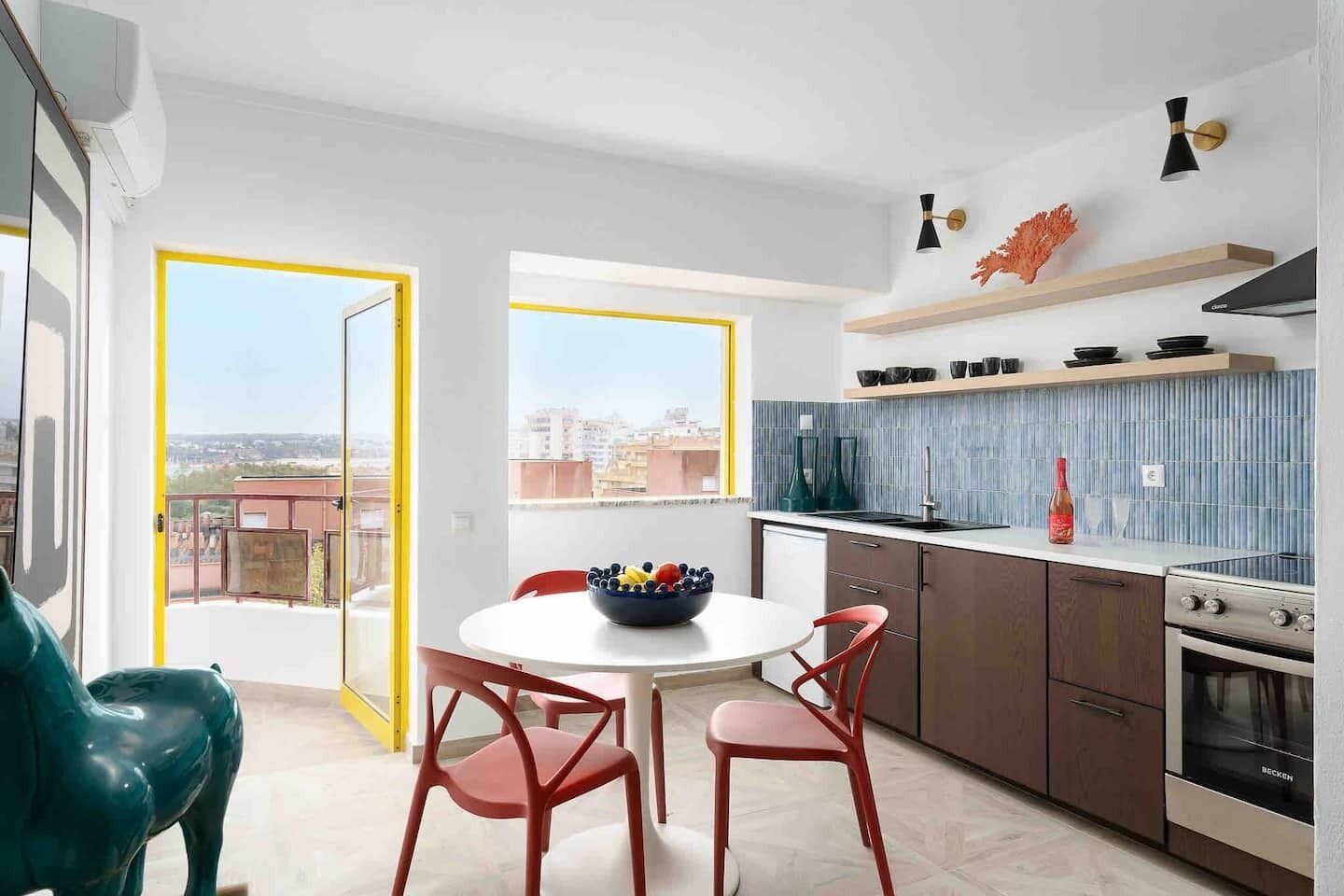 A modern kitchen with a dining area, featuring a round table with chairs, a bowl of fruit, and a view of the city through large windows.