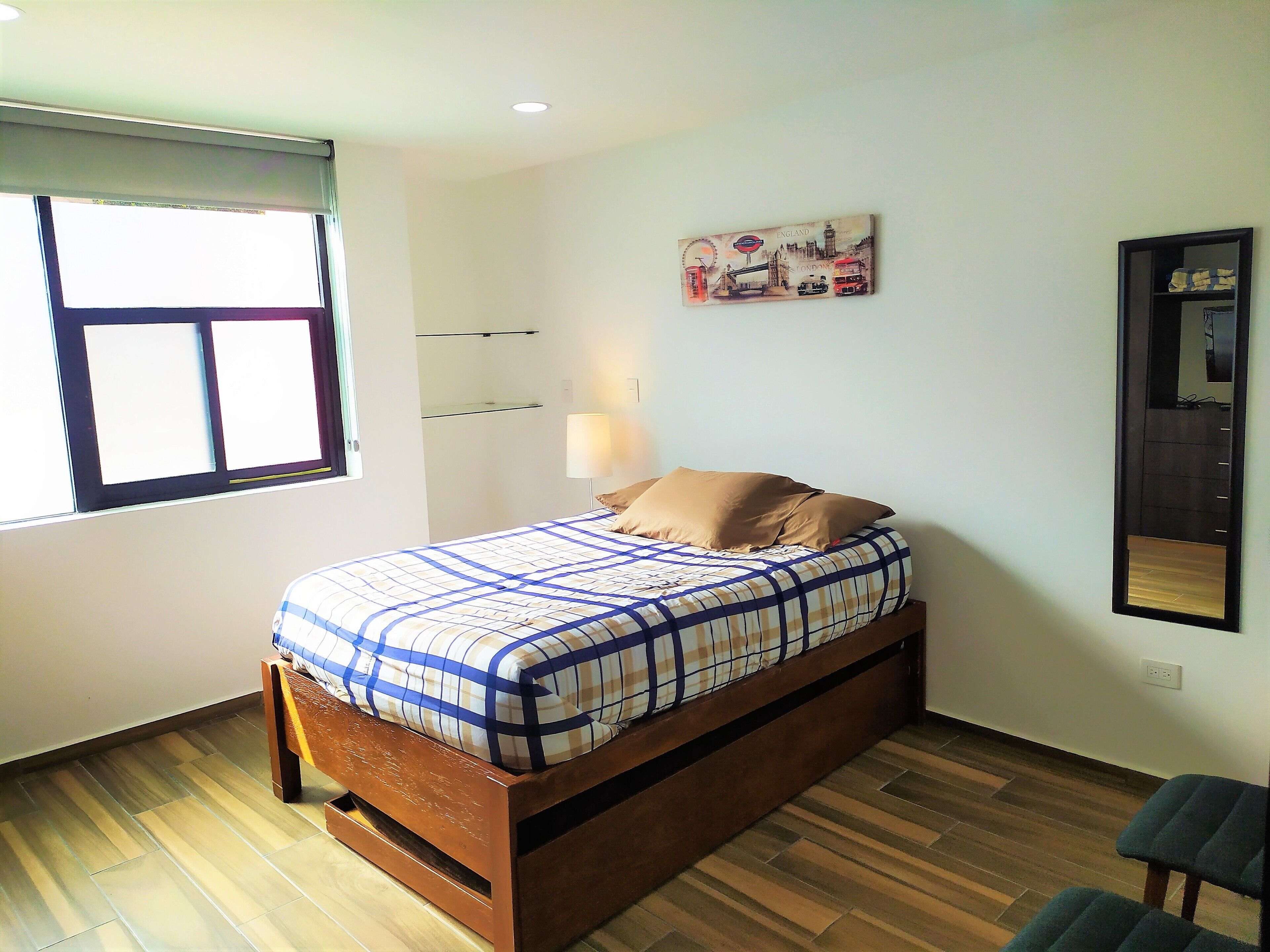 Un lit simple avec une couvre-lit à carreaux, une lampe de chevet, un miroir fixé au mur et une fenêtre avec des stores.