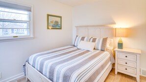 Un dormitorio con cama, mesita de noche, lámpara, ventana y un cuadro en la pared.