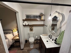 Une petite salle de bain comprenant des toilettes, un lavabo et un miroir avec un cadre décoratif. On y trouve des étagères au-dessus des toilettes et une petite fenêtre.