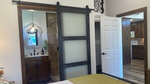 Une chambre à coucher comprenant un lit, une commode, un miroir et une porte coulissante donnant sur la cuisine.