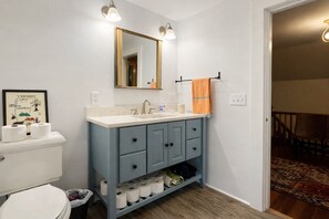 A bathroom with a white toilet, a blue vanity with a mirror, and a shelf with toilet paper. - Charming Chester Farmhouse near Near VT Slopes (Chester)