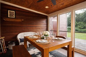 A wooden dining area with a table set for four, a wicker chair, and a view of the outdoors. - Charming Chester Farmhouse near Near VT Slopes (Chester)