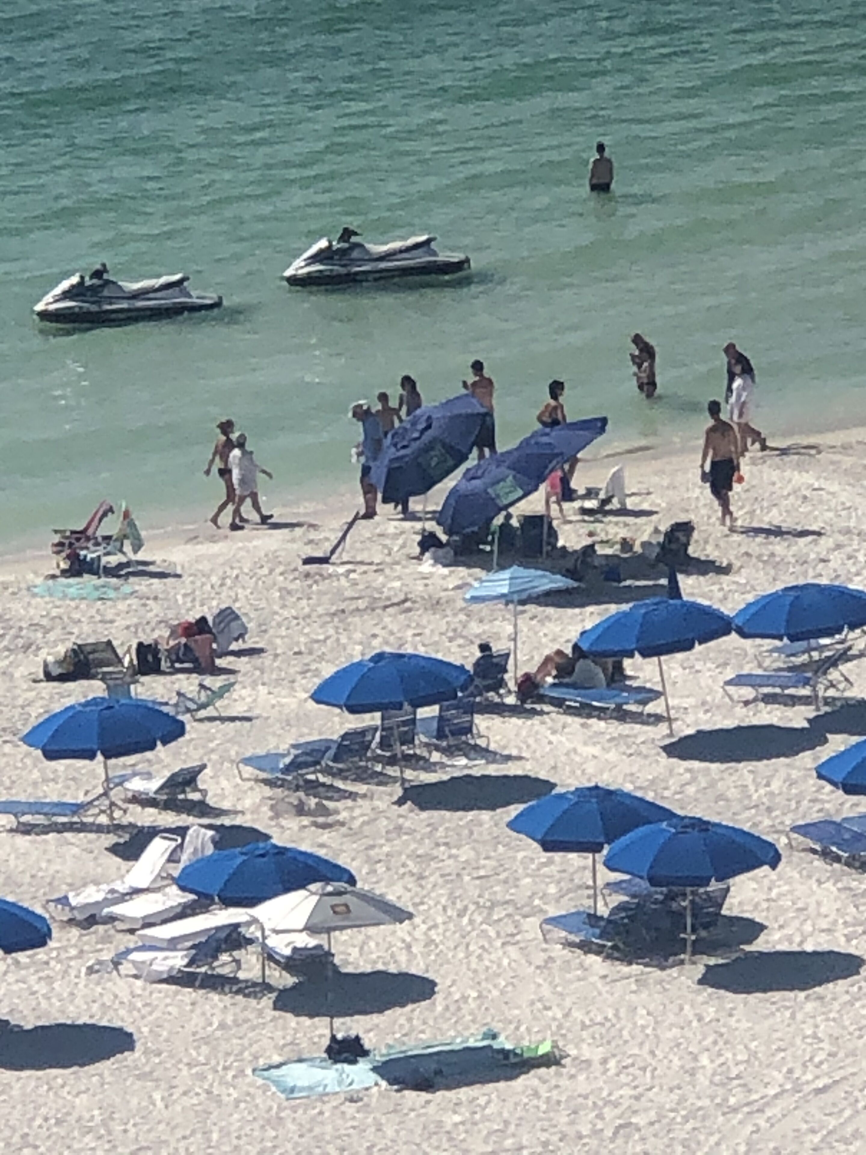 Una playa con múltiples sombrillas azules y sillas de descanso, un jet ski y gente caminando por la orilla.