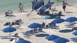 Ein Strand mit mehreren blauen Sonnenschirmen und Liegestühlen, einem Jetski sowie Menschen, die am Ufer entlanggehen.