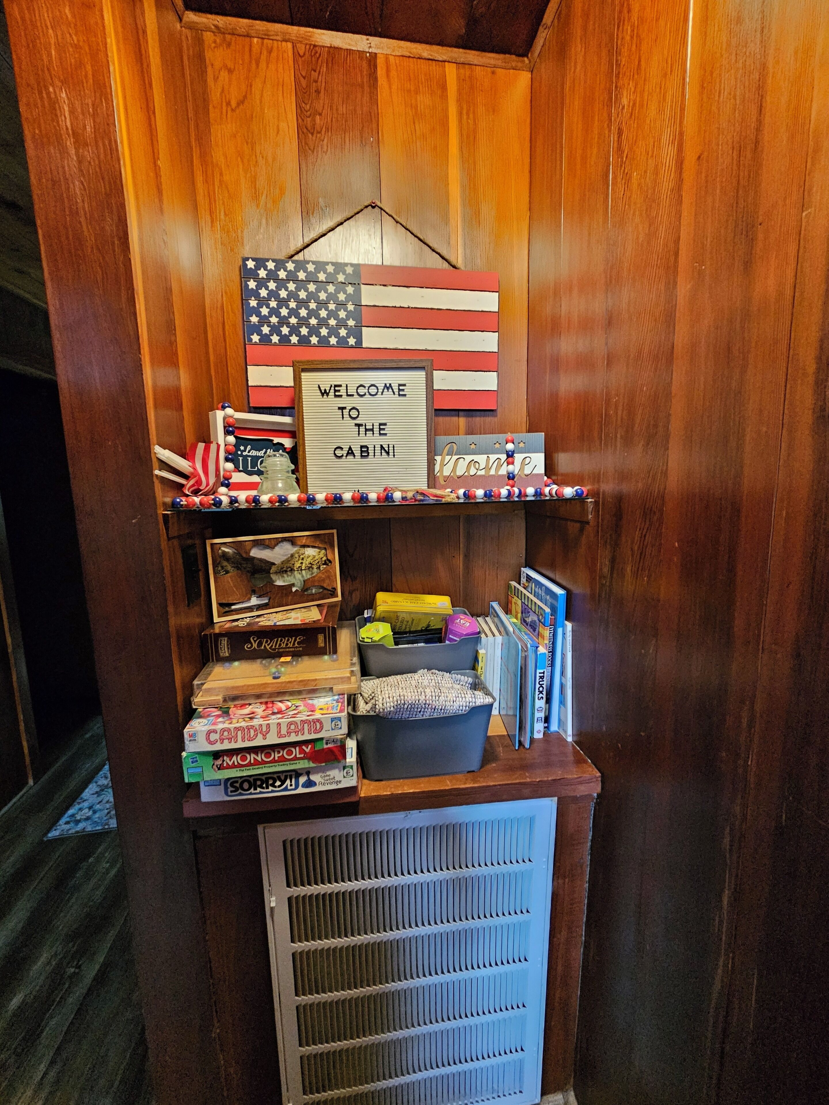 Interior de una cabaña de madera con un cartel de bienvenida, bandera estadounidense y estantes con juegos de mesa y aperitivos.