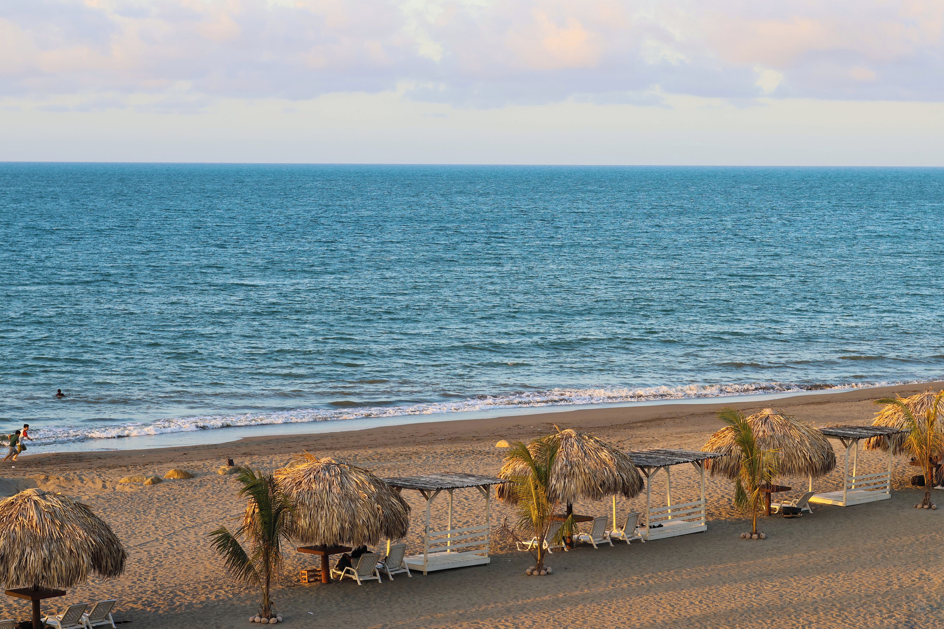 Una playa con sombrillas de techo de paja y sillones de descanso, una orilla arenosa y el océano.