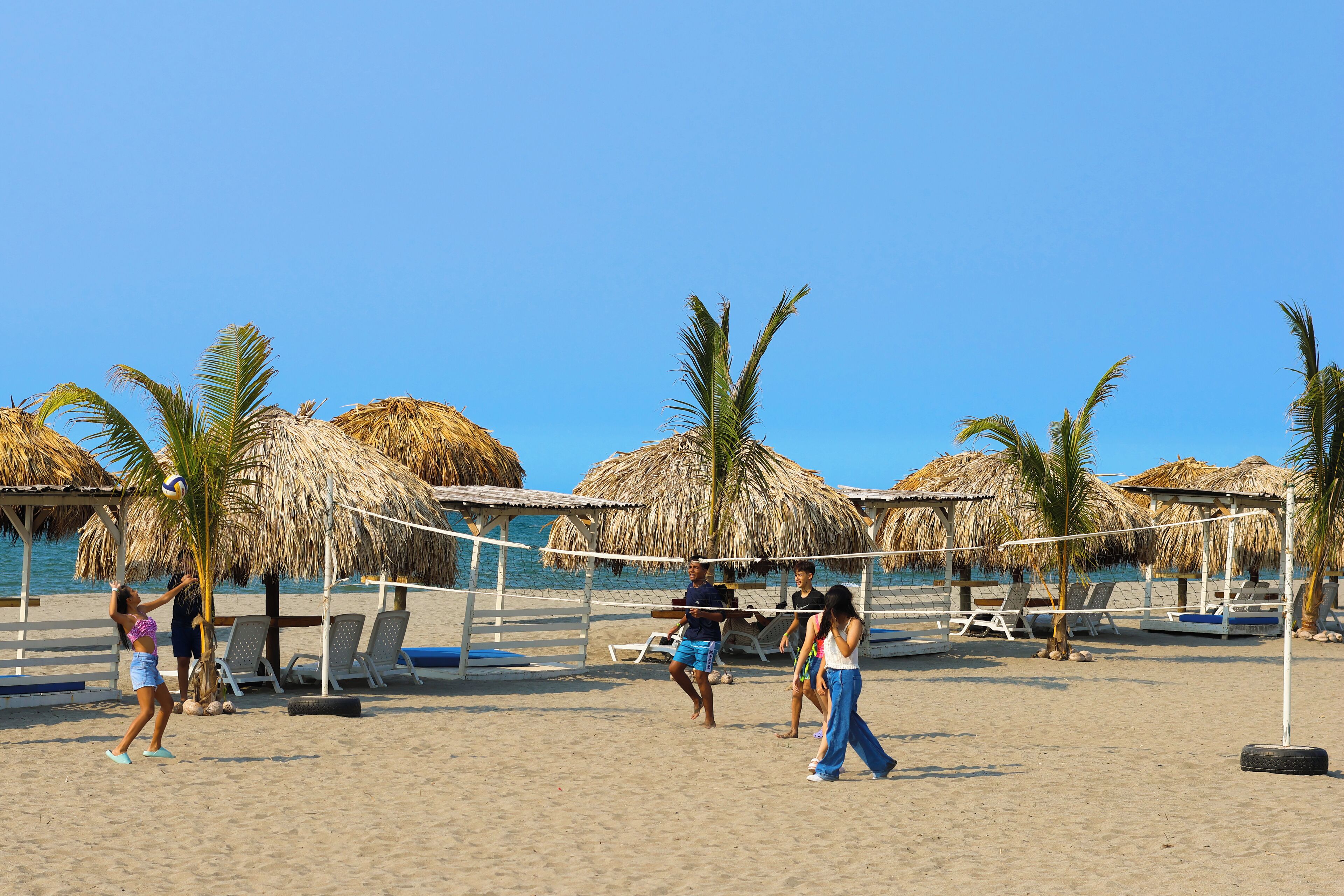 Una cancha de voleibol de playa con red, palmeras y estructuras de techo de paja.