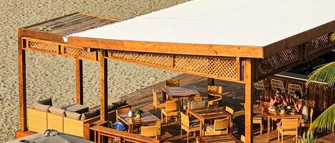 A beachside restaurant with outdoor seating, a covered area, and a view of the ocean.